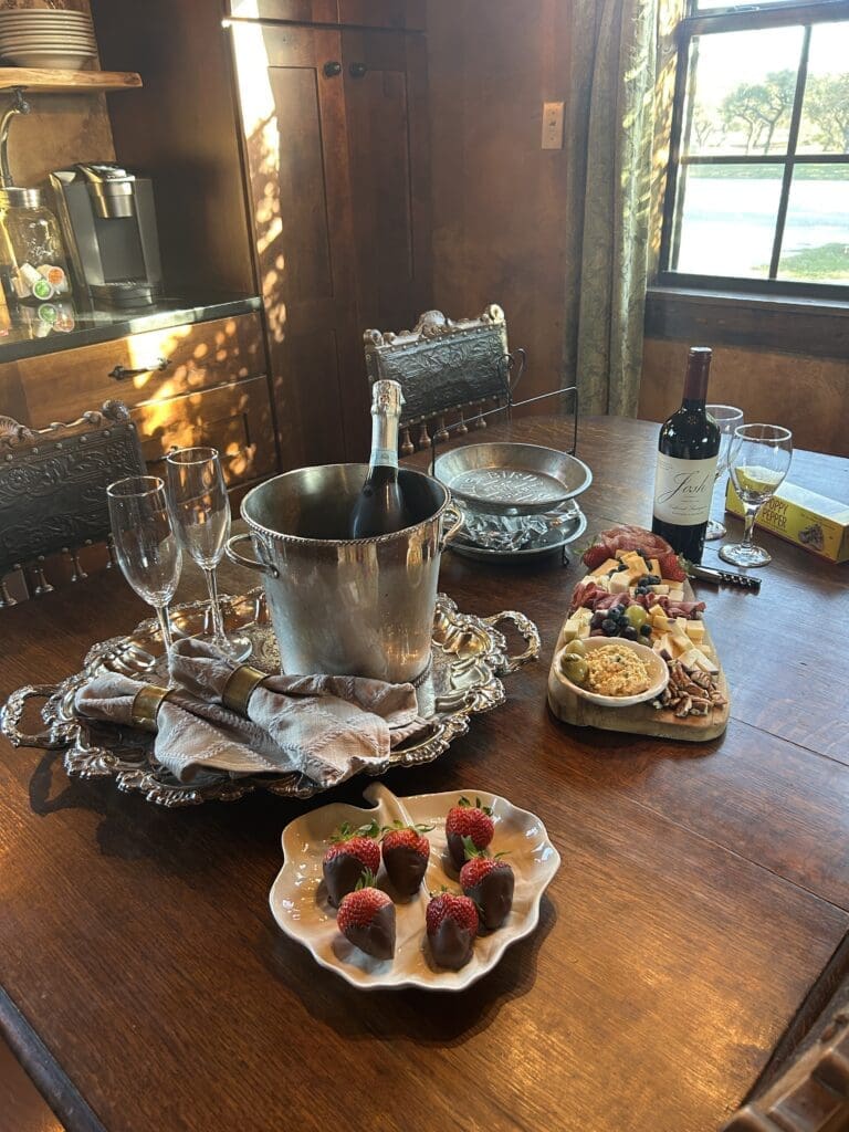 IMG_8254 - Inn at Sunset Mill Ranch Champagne in a silver bucket sits on a wooden dining table beside a cheese board with chocolate-dipped strawberries and a bottle of wine.