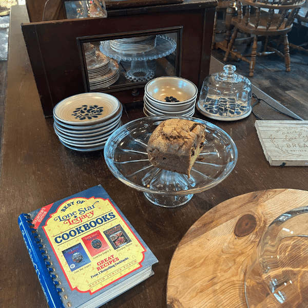 Loaf on a glass cake stand on a wooden table with stacked plates and a cookbook nearby in a cozy kitchen display.