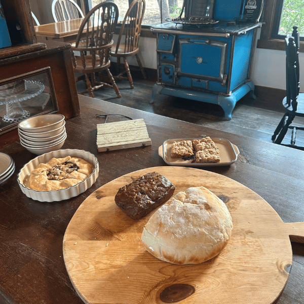 Round wooden board with two loaves (one light, one dark) and pastries on a rustic kitchen table; blue vintage stove in the background.