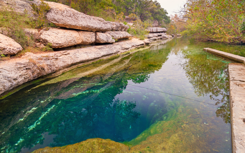 Clear turquoise pool with rocky limestone walls and reflections of trees; a wooden dock runs along the right edge.