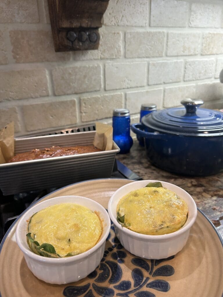Two individual ramekins of baked egg dish on a decorative plate in a kitchen, with a blue Dutch oven and a baking pan in the background.